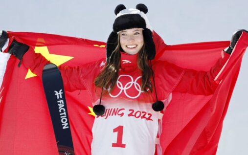 Eileen Gu holding her skis and smiling at the bottom of the slope after a freestyle skiing run