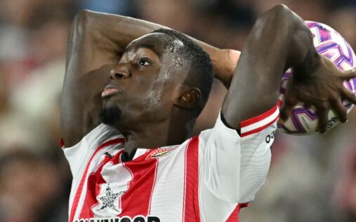 A football referee checking their watch while a player prepares to take a throw-in on the touchline