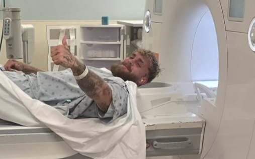 Jake Paul sitting in a hospital bed with a bandaged jaw alongside Jutta Leerdam