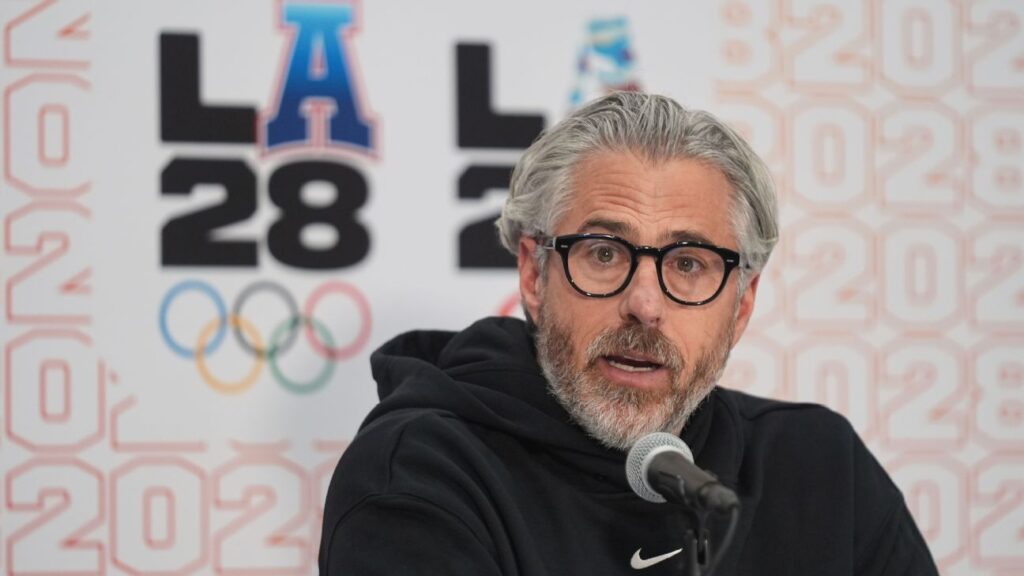 Casey Wasserman speaking at a podium with the LA 2028 Olympic logo in the background