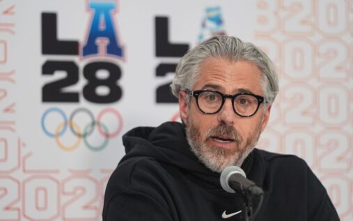 Casey Wasserman speaking at a podium with the LA 2028 Olympic logo in the background