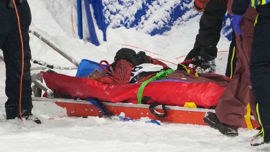 Close up of Mark McMorris wearing a snowboard helmet and goggles, looking determined against a snowy mountain backdrop