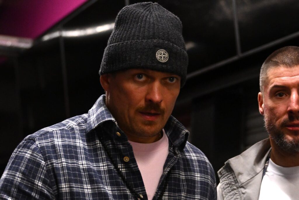 Oleksandr Usyk celebrating in the ring with heavyweight championship belts draped over his shoulders