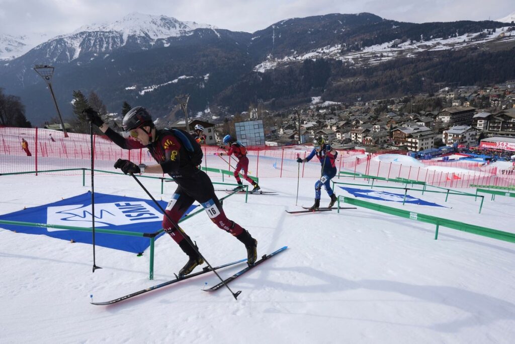 A ski mountaineering athlete ascending a steep snowy slope with skis attached to their backpack during a competition