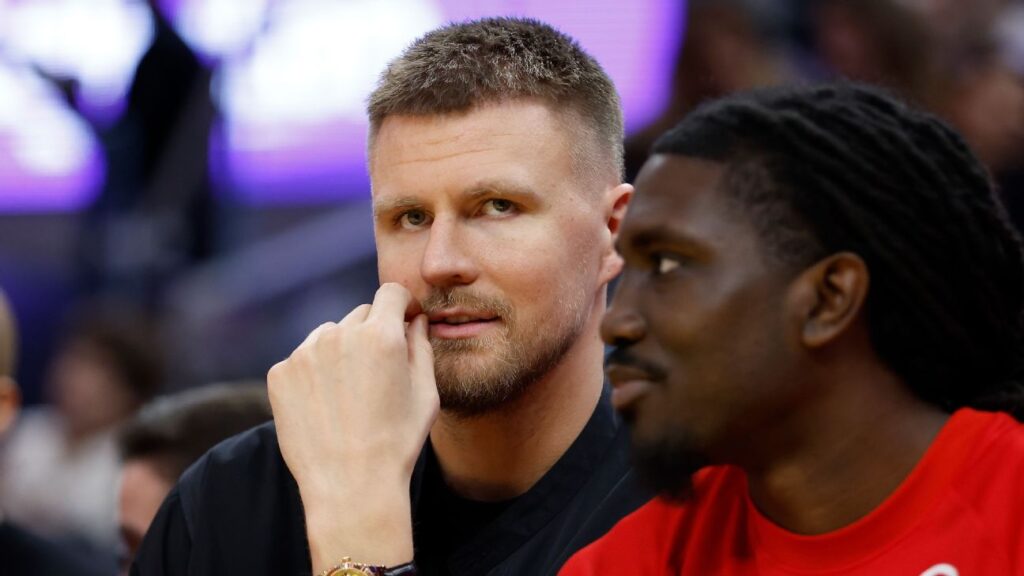 Stephen Curry watching from the bench while Kristaps Porzingis warms up in Golden State Warriors training gear