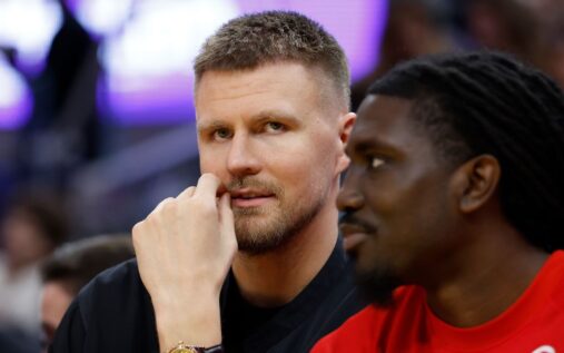 Stephen Curry watching from the bench while Kristaps Porzingis warms up in Golden State Warriors training gear