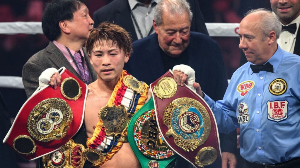 Naoya Inoue standing in the boxing ring holding his championship belts