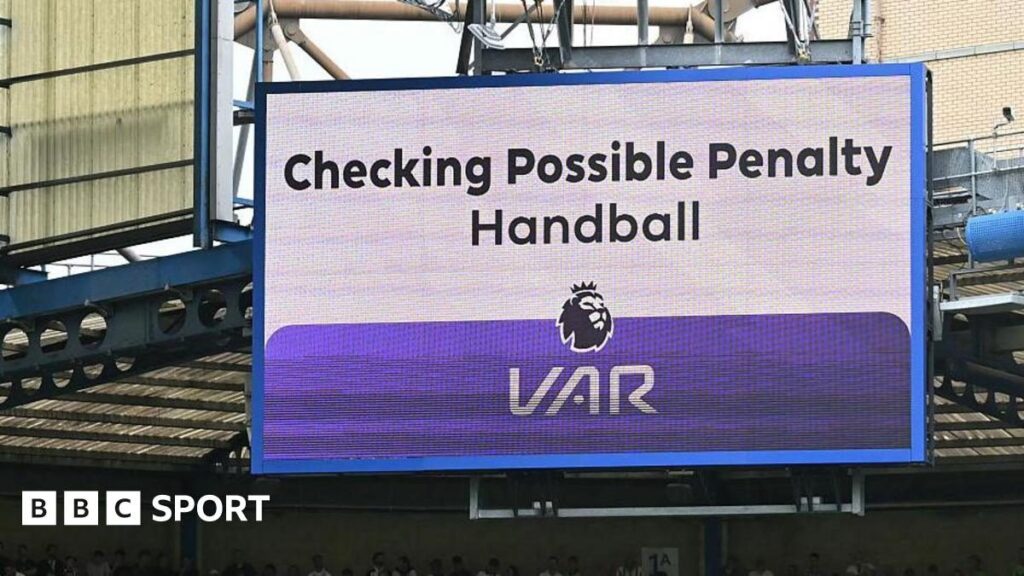 A referee reviewing a pitchside VAR monitor during a Premier League match, reflecting fan frustration with the technology.