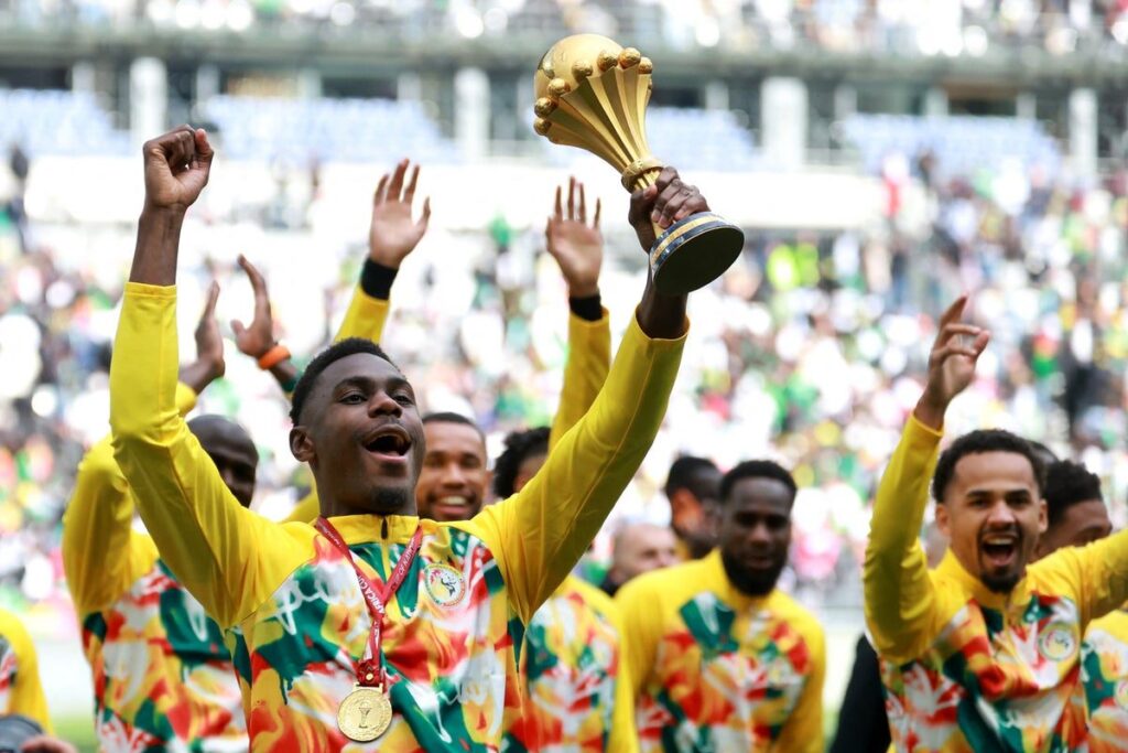 Senegal football players lifting and celebrating with the Africa Cup of Nations trophy in front of fans at Stade de France.