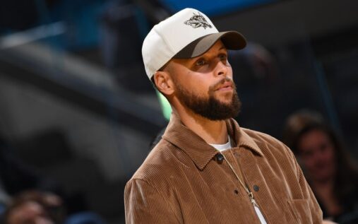 Stephen Curry sitting on the Golden State Warriors bench wearing warm-up gear