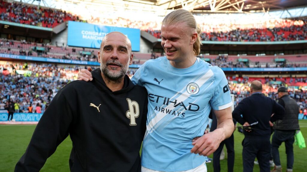 Pep Guardiola smiling on the Manchester City touchline wearing a casual sweatshirt