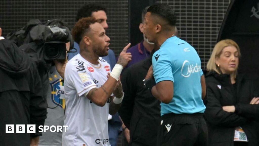 Neymar looking furious on the touchline while holding up a substitution slip to a camera