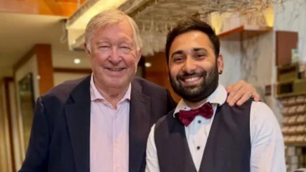 Sir Alex Ferguson smiling warmly alongside a supporter in a restaurant following his recent recovery